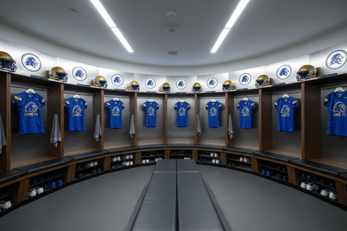 Professional sports locker room bay with 12 open athletic lockers, uniforms hanging, team logos on every back panel, wall-mounted bench in foreground